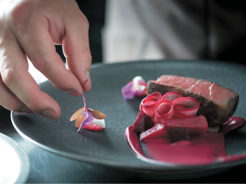 シェフの手が丁寧に仕上げる、黒いプレートの肉料理。鮮やかなソースと花びらの装飾が添えられ、一皿に込められたこだわりが伝わります。